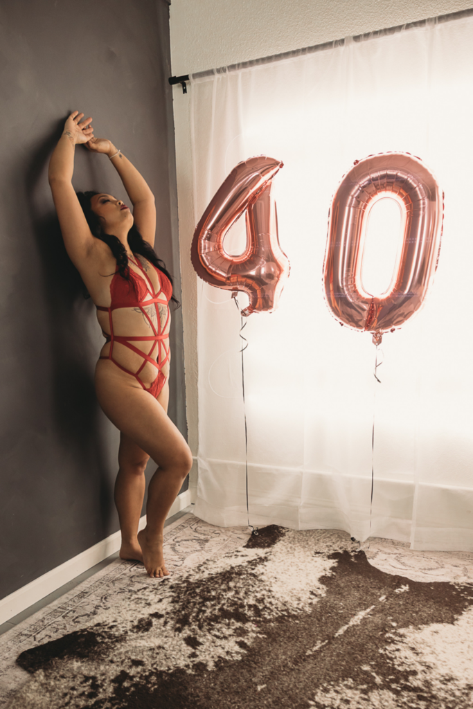 Boudoir over 40 session highlighting confidence, self-acceptance, and feminine empowerment as a woman embraces her beauty in a red lingerie portrait with rose gold birthday balloons.