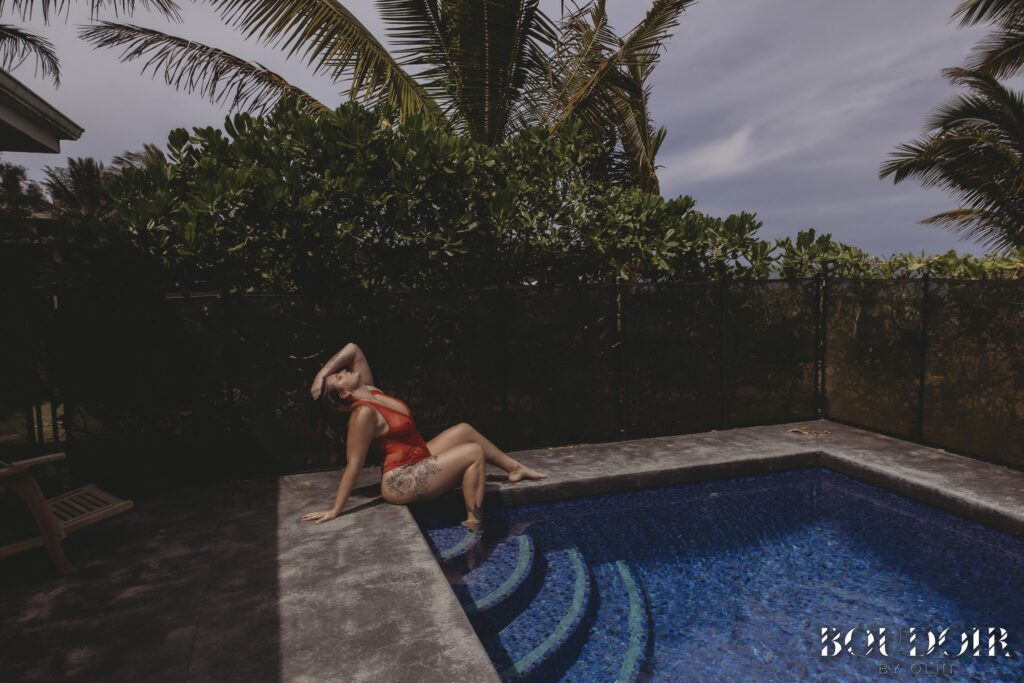 A woman reclines on the edge of a pool in a red lingerie bodysuit, her head tilted back and one arm draped over her forehead. Lush tropical plants and palm trees surround the pool, with sunlight casting soft shadows. This image exemplifies women's portrait photography Hawaii, capturing confidence, elegance, and natural beauty in an outdoor setting.