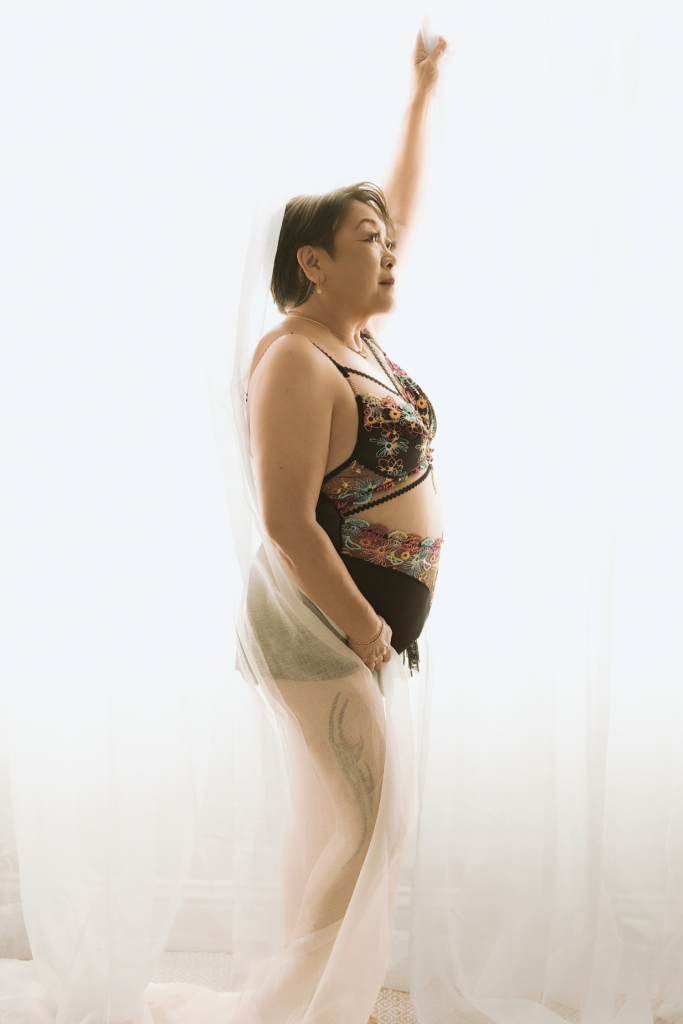 Older woman posing beside sheer white curtains in embroidered lingerie and a flowing sheer wrap during a softly backlit Sacramento boudoir portrait session.