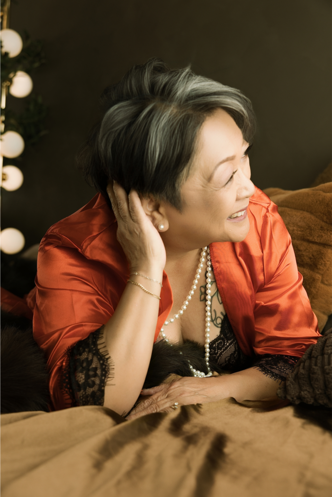 Older woman smiling in a red satin robe with black lace trim and pearl jewelry during an elegant Sacramento boudoir photography experience.